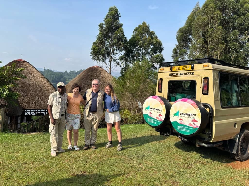 Viaggiatori e la loro guida sul prato di un lodge con banda dal tetto di paglia, accanto al loro veicolo da safari
